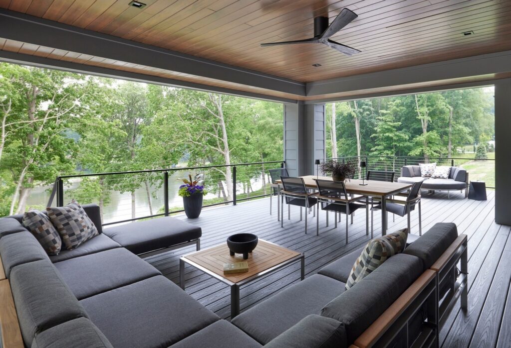 An outdoor patio with gray lounge seating, a dining table, and a view of a lush green forest and a body of water.