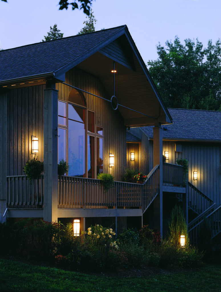 A house with a large front porch illuminated by various outdoor wall lights and ground lights, set against a dark blue evening sky.