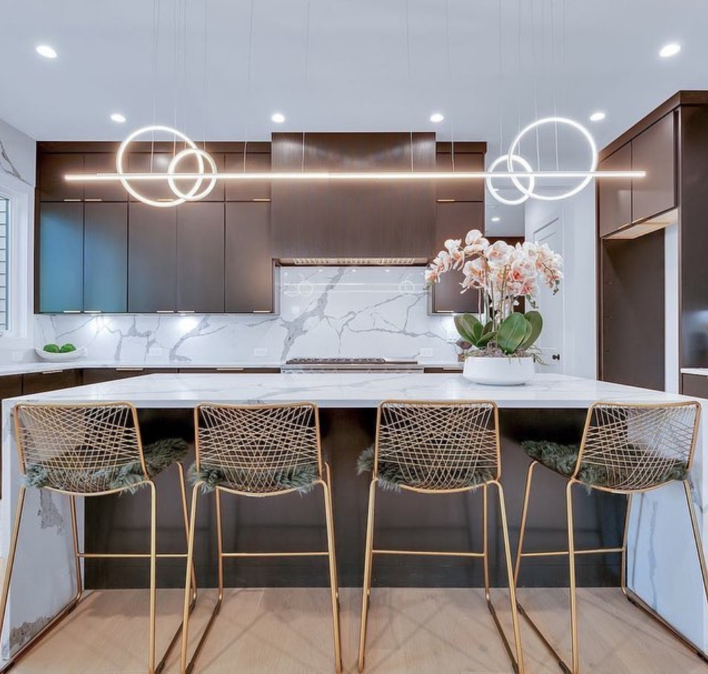 A modern kitchen features dark cabinetry, a marble backsplash and island, and unique circular and linear pendant lights.