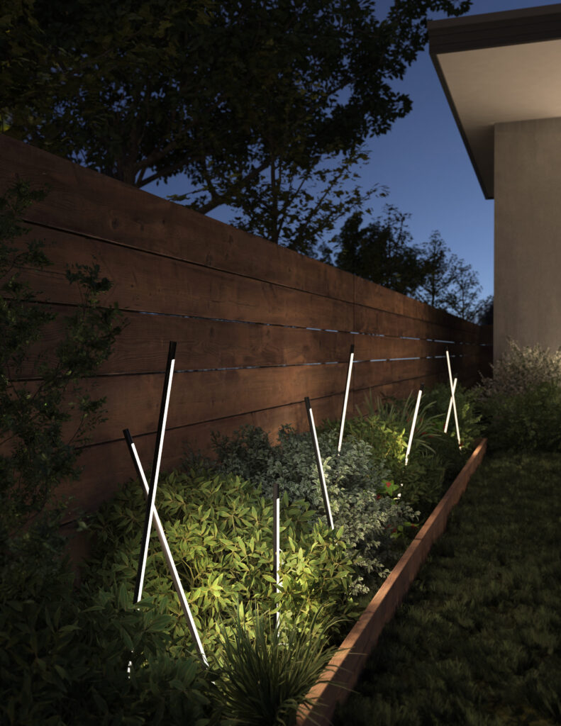A modern garden at dusk, featuring slim, vertical uplights illuminating lush green plants along a wooden fence.