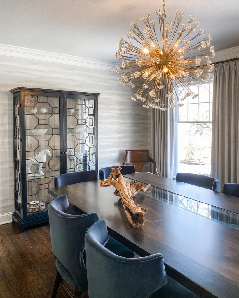 A modern dining room features a dark wood table with blue velvet chairs, a large sputnik chandelier, and a display cabinet.