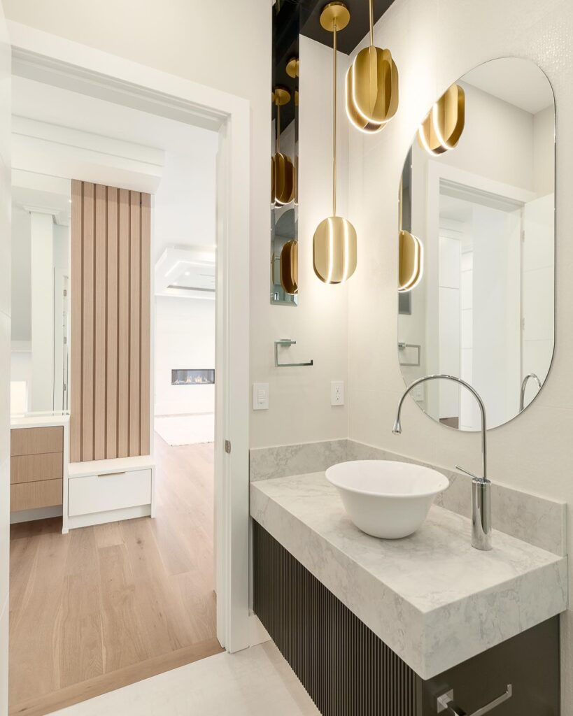 A contemporary bathroom with a vessel sink, marble countertop, gold pendant lights, and an oval mirror.
